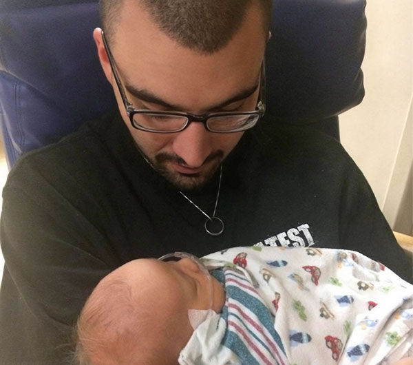Joey looking down holding his newborn son in his arms, son wrapped in a hospital blanket